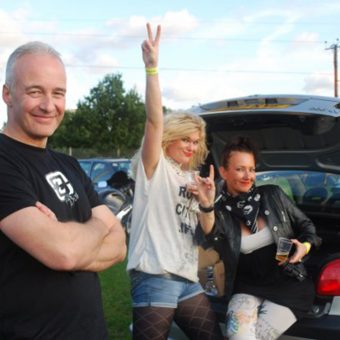 Neil on the left, standing grinning with his arms folded. In the background, an attractive blonde girl smiles and gives the peace sign next to her friend, an attractive brunette, who sits in the boot of their open car giving the rock salute.