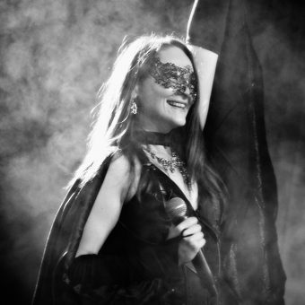 Ann wearing batwings and a Halloween mask, smiling and raising her left hand while holding her mic in her right hand. Black and white.