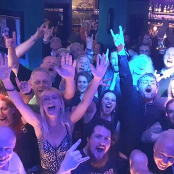 The crowd at the Pub, Lancaster, as seen by the band from the stage. Everyone is cheering, smiling and waving their arms, and some are throwing the rock horns. Band members, John, Pete, Aaron and Neil have also photo-bombed the pic.