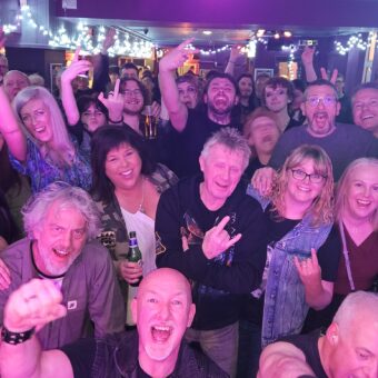 The crowd at Grand Central, Manchester, as seen from the stage. Everyone is smiling, cheering and throwing the horns. Guitarists John and Neil have also photo-bombed the pic.